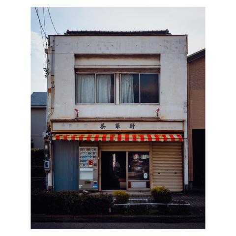 A two-story concrete building with weathered white walls stands modestly between more modern neighbors. The upper windows are curtained in off-white, just sheer enough to hint at the life within. Below, a striped red-and-yellow awning gives a faint carnival energy to what is otherwise a portrait of stillness. A cigarette vending machine beside the door lends a timestamp.