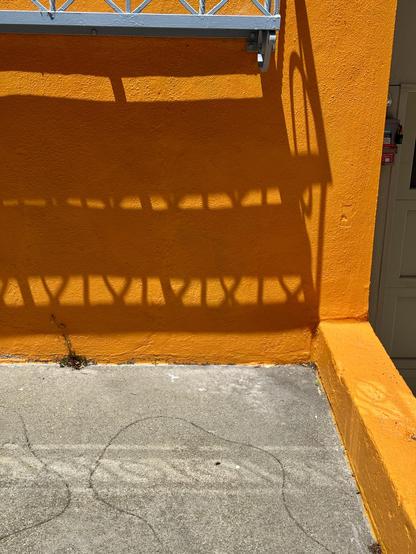 The side of a house with orange walls and a concrete driveway. There is a shelf under the front windows that is casting a shadow on the wall beneath it. This shadow was used as inspiration for my drawing of several orks, but this is the original photo. 