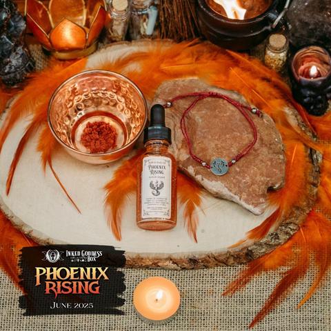 A small hammered copper bowl contains a pinch of orange powder. A small clear vial with a black eyedropper top contains orange-ish liquid. An adjustable bracelet made with red knotted cord and a silver phoenix charm sits on a red rock. These are three of the items in the exclusive June 2025 "Phoenix Rising" Inked Goddess Creations Box.