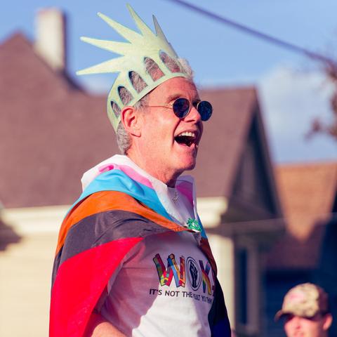 Man wears statue of liberty cap and dark round glasses and a multi colored case and a shirt that says WOKE: IT'S NOT THE INSULT THEY THINK IT IS