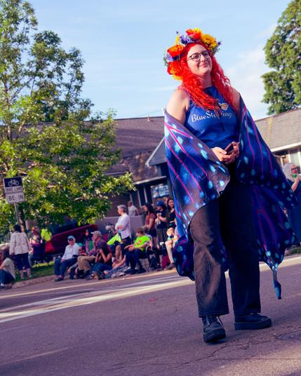 Woman with orange hair with flowers and a butterfly cape and blue top that says Bluestockings has dark pants and boots in the middle of a street with people sitting on the far side with a short building behind and a few looming trees