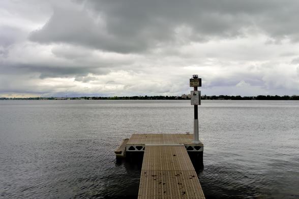 A landscape scene dominated by water and sky under an overcast sky. A weathered dock extends from the foreground into the calm water, leading the eye towards a distant shoreline. A signpost stands on the dock, displaying a caution sign about shallow water and no diving. The sky is filled with heavy, textured clouds, casting a muted light over the scene. The overall color palette is cool and subdued, with various shades of gray and subtle hints of green and brown in the distant vegetation. The photograph evokes a sense of tranquility and solitude.