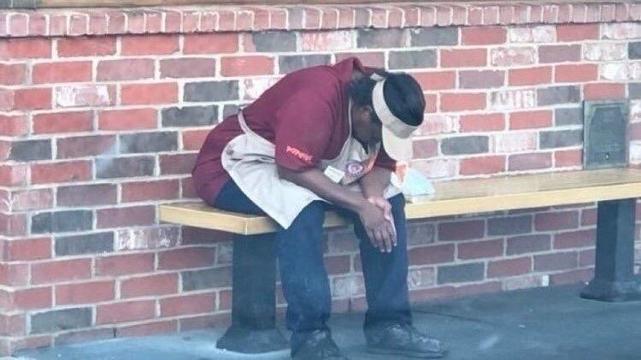 Popeyes Woman meme. 
A worker in a red shirt and apron sits on a bench, leaning forward with their head down, looking tired or distressed. The backdrop features a brick wall.