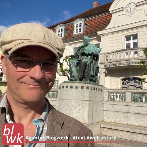 Autor Oliver Hübner mit Kappe am Fritz-Reuter-Literaturmuseum in Stavenhagen, Skulptur von Fritz Reuter