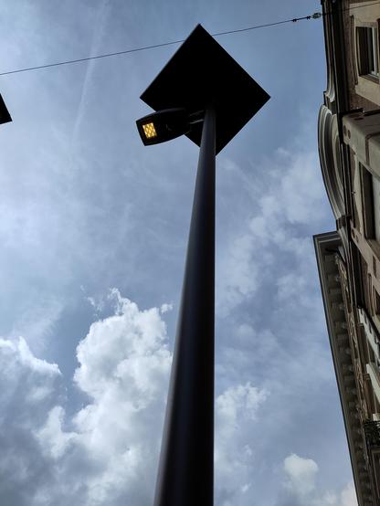 Zu sehen ist eine Straßenlaterne aus stärker Untersicht. Sie hat einen LED-Leuchtkörper, der aktiv ist, sowohl es taghell ist. Direkt über dem Leuchtkörper befindet sich ein quadratisches Solarpanel. Am Bildrand ist eine Hausfassade angeschnitten. Am Himmel ist eine leichte Bewölkung erkennbar.