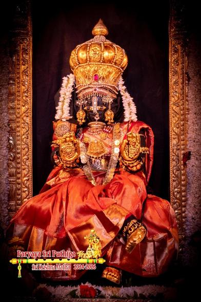 Idol of Hindu diety Krishna dressed in red as Lakshmi (feminine) sitting cross-legged (right leg over left), wearing gold crown, gold shoes, and several gold ornaments. Two small strings of white flowers, one on either side of Krishna, fall down from the side of the crown to Krishna's shoulders. The sides of a gold frame around Krishna are visible. Part of a thick white flower garland attached to the frame is also visible.