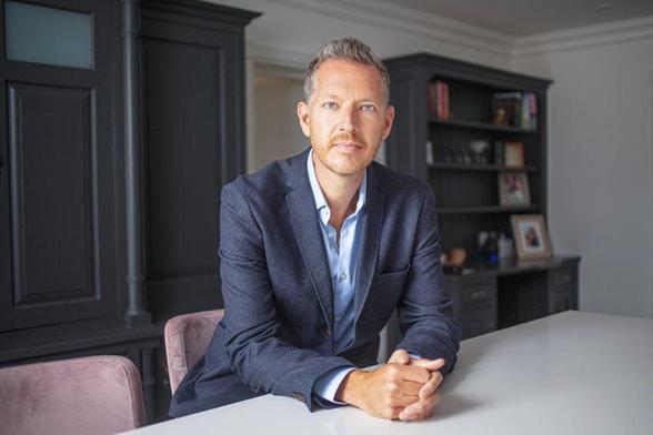 A photo of Nick leaning on an office desk, wearing a grey suit jacket and a light blue shirt.
