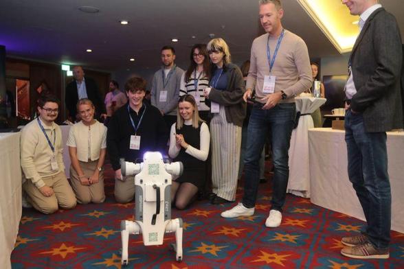 A grey and white robot dog doing a hand-stand in a crowded conference room.