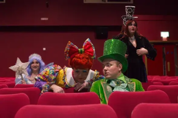 A group of people dressed in colorful, theatrical costumes sits in a theater with red seats. One person wears a glittery green top hat and green outfit, another has a large red wig with a big bow and a multicolored dress, a third is dressed as a fairy with a crown and star wand, and a fourth wears a black outfit with a tall, patterned hat. They appear to be watching a performance, with some leaning forward in their seats. The theater has red walls and a few visible exits in the background.