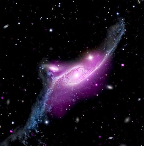 Centered in the frame, NGC 6872 is a large barred spiral galaxy with two elongated arms that stretch toward the upper right and lower left. Near the white dot at the heart of the galaxy, a cloud of neon purple tints the arms, which appear steel blue at the tips. The purple represents hot gas. Just to the upper left of NGC 6872 is a second spiral galaxy. Its spiraling arms are much smaller, but the bright white dot at its core is quite large. Some of the blue matter and gas from NGC 6872’s lower arm appears to be floating toward the smaller galaxy. The background of space is black, dotted with distant pinpoints of light in variety of colours.