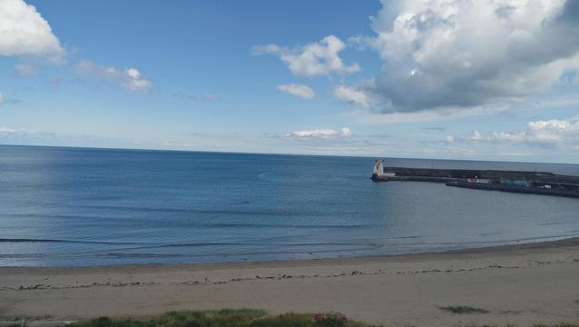 A view from Balbriggan train station, the beach can be in the foreground with old lighthouse at the end of the harbour wall to the right of the image. The sea is calm with a swimmer doing lengths between two buoys. The sky overhead is clear blue in the centre with white clouds to the right and left.