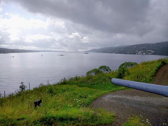 View over the Oslo fiord. Facing southwards. To the left in the picture, the eastern side of the fiord, Drøbak, to the right Storsand on the western side. In the foreground a grassy slope and the barrel of an old cannon aimed towards the waterway in the fiord. 