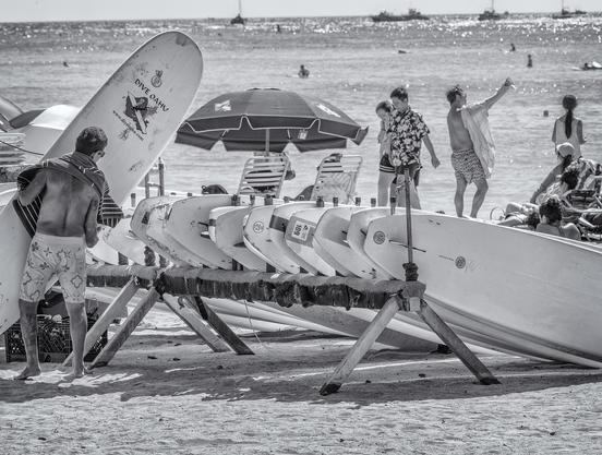 Man returns surf board to a rack of rental boards. In the background is Waikiki Beach with many people on the sand and in the water.