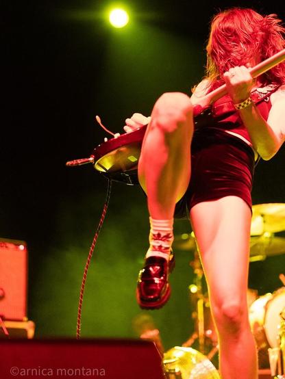 A female musician performs on stage, playing an electric guitar. She has long, flowing hair and is wearing a black outfit and platform shoes. The scene is illuminated with vibrant green and yellow stage lights, creating a dynamic atmosphere.