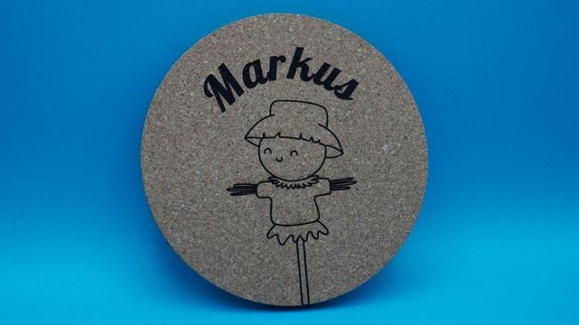 Runder Korkuntersetzer mit einem gravierten Motiv eines lächelnden Strohmanns und dem Namen „Markus“ darüber.