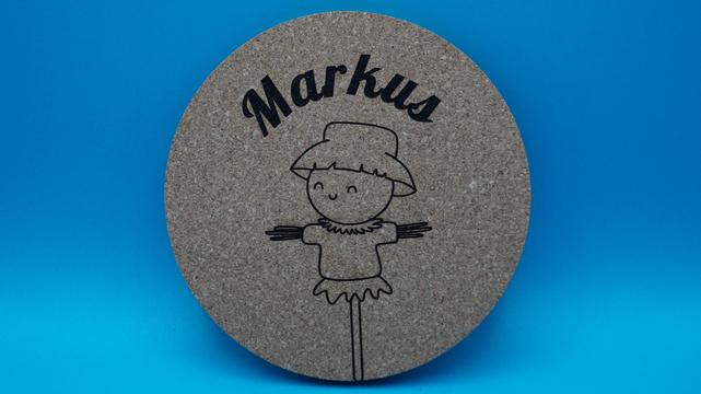 Korkuntersetzer mit Strohmann-Figur und personalisiertem Namen „Markus“, leicht angewinkelt dargestellt, vor blauem Hintergrund.