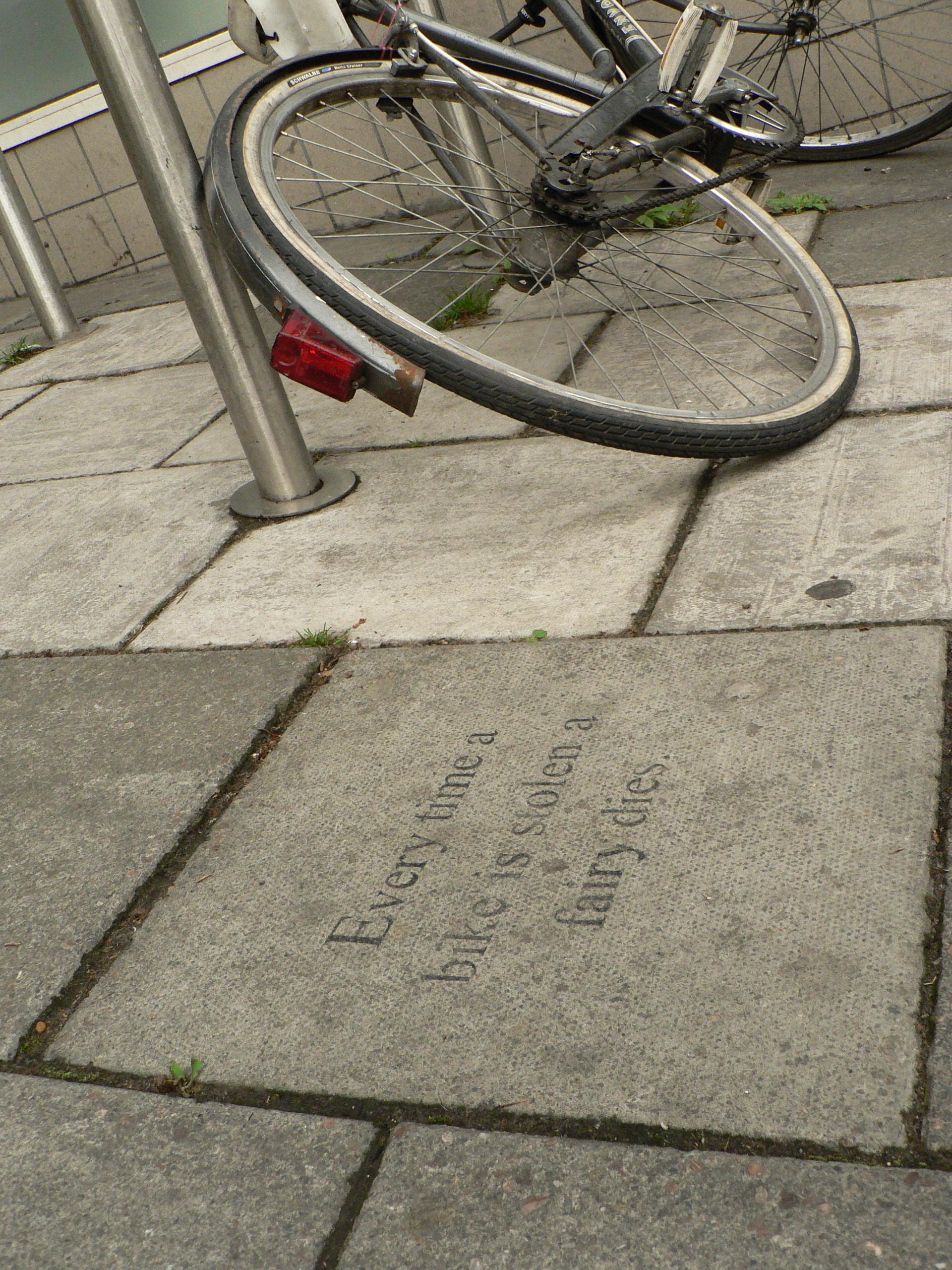 Dated photo of part of a broken bike still tied to a bike stand. In the foreground, a stencil reads "Every time a bike is stolen a fairy dies" in a neat, serif font.