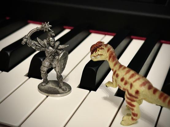 A green Dilophosaurus with brown spots faces off with a small pewter figure of what I think is a human warrior dressed in elaborate plate armour and holding a mace above his head with his right hand while bracing a shield in the left. Both stand on a piano keyboard.
