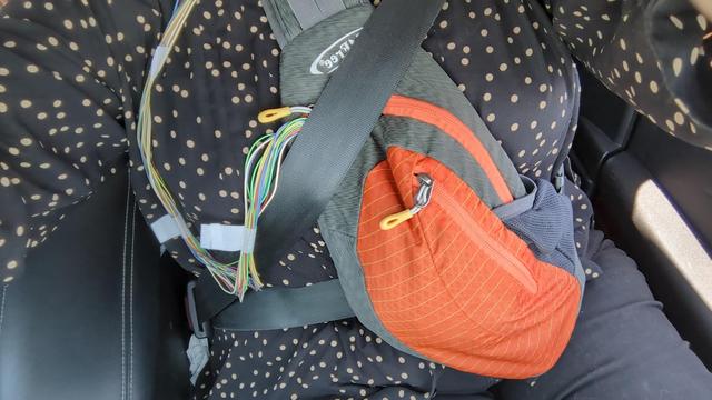 A photo of Ivy's front of body - she's seatbelted in a passenger seat. A front bag, bigger than a fanny pack but smaller than a backpack, is strapped to her body with medical lead wires snaking from the top of the screen where her head is and down into the bag