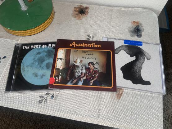 An inage showing a table with three music CDs. From left to right, the CDs include "The Best of R.E.M. 1988–2003” by R.E.M., “Here Comes the stunts” by AWOLNATION, and “Fragrant World” by Yeasayer. The R.E.M. and Yeasayer discs are in jewel cases, while the AWOLNATION disc is in a paper sleeve.