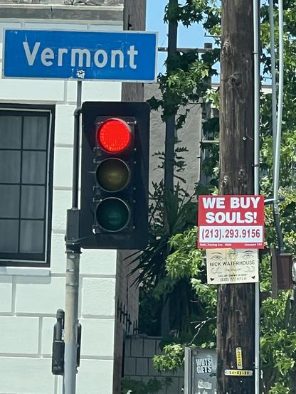 The image features a traffic light showing red, a street sign for "Vermont," and a large advertisement that says "WE BUY SOULS!" with a phone number. There are also plants nearby and a utility pole with additional signage.
