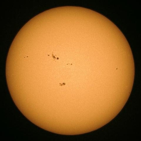 A close-up image of the Sun, displaying its bright orange surface with several dark sunspots visible.