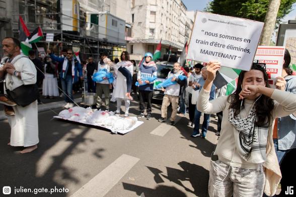 Nous sommes boulevard Marguerite de Rochechouart. Des manifestants mettent en scène, à travers une scénographie poignante, la situation actuelle de la population de Gaza. Un premier manifestant, pieds nus, attaché à une corde, tire une planche sur laquelle reposent cinq corps enveloppés dans des draps blancs, maculés de faux sang. Derrière lui, quatre autres manifestants prolongent le tableau. Le premier porte une chasuble « UNRWA » et tient dans ses bras le corps d’un enfant enveloppé dans un plastique bleu. À ses côtés, un manifestant déguisé en chirurgien porte un squelette vêtu d’un pantalon. À sa droite, un autre manifestant, équipé d’un casque bleu et d’un gilet pare-balles marqué « PRESS », tient lui aussi un corps d’enfant. Enfin, le dernier manifestant de la file en porte un également. Au premier plan, une jeune femme en pleurs brandit une pancarte : « Nous sommes en train d’être éliminés, MAINTENANT ». Depuis le début de l’offensive israélienne, le nombre de morts estimé dans la bande de Gaza dépasse les 60 000 personnes, et plus de 110 000 ont été blessées.