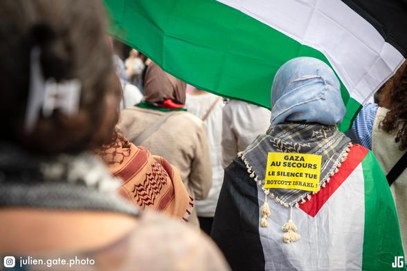 Le cortège arrive place Saint-Augustin. Une manifestante arbore un drapeau palestinien sur le dos. Autour de son cou, un keffieh, sur lequel est collé un autocollant : « Gaza au secours, le silence tue, boycott Israël ! ». Devant elle, un autre drapeau palestinien flotte dans le vent ; sa bande verte semble se prolonger visuellement sur celui qu’elle porte.