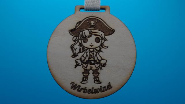 Holzmedaille mit Gravur eines Piratenmädchens und der Aufschrift „Wirbelwind“. Die Medaille hängt an einem weißen Baumwollband mit Klettverschluss – vor blauem Hintergrund.
