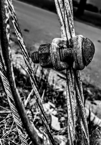 A close-up black and white photograph showing rusty, weathered steel cables being held together with a U-bolt secured by a nut and fastener. The metal parts show signs of age and corrosion. Background is blurred road.