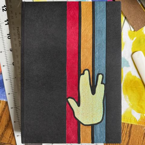 Handmade book with a black cover, the three stripes in the colors of star trek the original series uniforms (red, gold, and blue) with a green “Vulcan salute” hand print. 