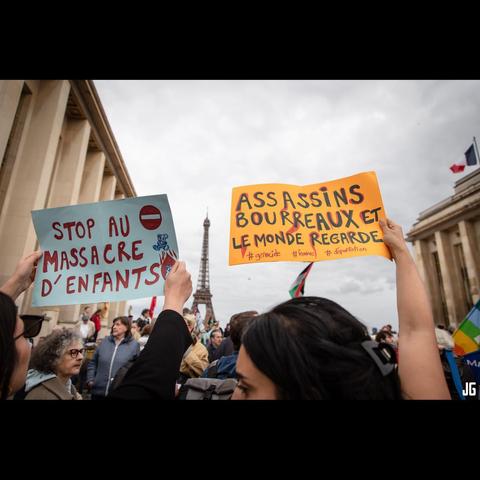 Deux manifestantes tiennent des pancartes sur lesquelles sont inscrits les messages suivants : « Stop au massacre d’enfants » et « Assassins et bourreaux, le monde regarde ». En arrière-plan, la tour Eiffel et un drapeau français flottant sur un bâtiment sont visibles.