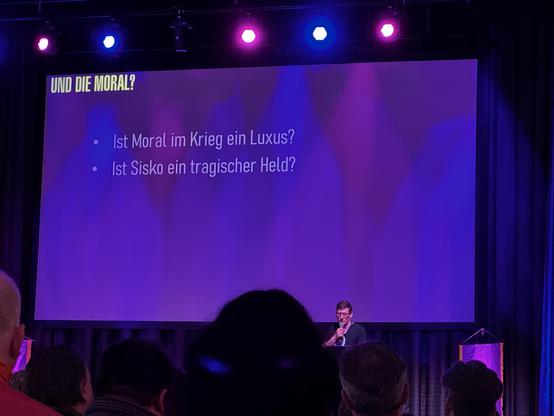 The image shows a speaker at an event, addressing an audience. A projection screen displays two questions in German: "Ist Moral im Krieg ein Luxus?" and "Ist Sisko ein tragischer Held?" The setting has vibrant colored lighting.