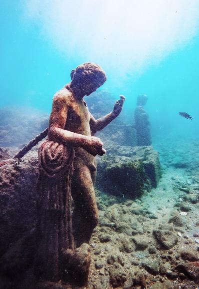 A marble statue of Dionysos under water, seen from the side. He is leaning on a plinth with a himation draped over it. In the background, we can see a fish and another statue.