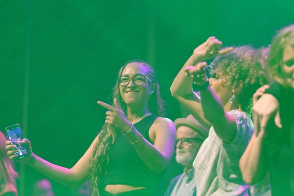 A group of people enjoying a lively performance on stage, with one woman in the foreground smiling, holding a phone, and dancing. The scene is illuminated with green lighting, creating a vibrant atmosphere.