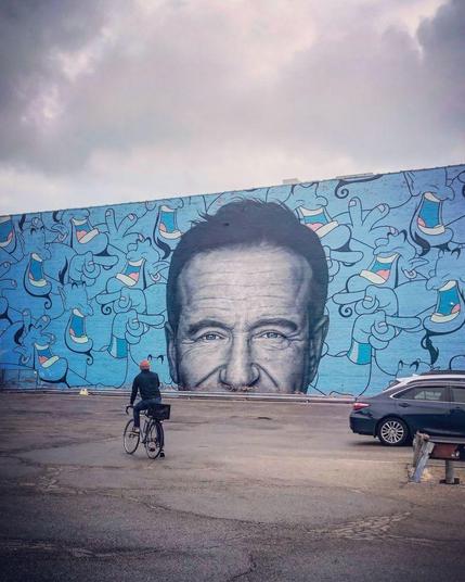 Mural of Robin Williams surrounded by various illustrations of the genie character he portrayed/voiced in the Disney movie, Aladdin. Situated in front of the mural is a lone bicyclist, propped up on one leg as they straddle their bike.