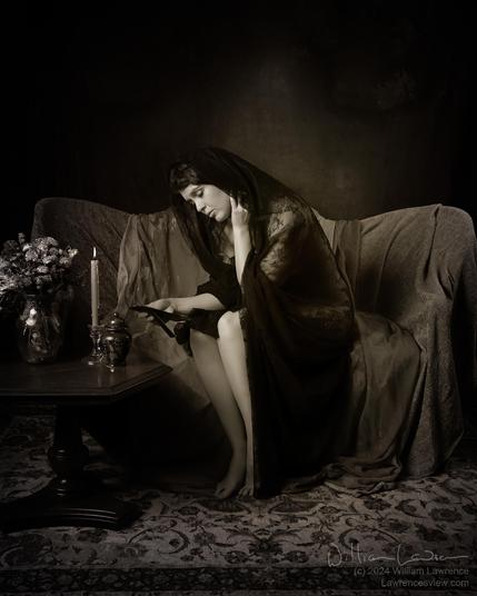 Model Jessa Ray Muse poses sitting on a draped settee, herself draped in sheer black fabric.  She gazes sadly at a photography, with an urn, a melting candle, and a vase of flowers off to the side.