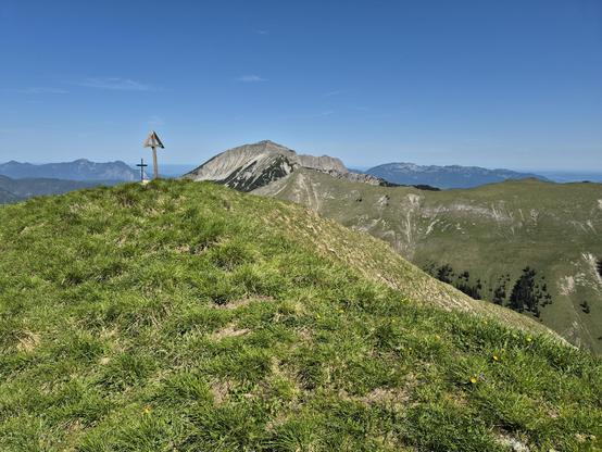 A scenic view from a grassy hilltop with a wooden cross and a triangular structure marking the summit. In the background, rugged mountains with rocky peaks and valleys stretch under a clear blue sky. The landscape includes patches of green grass and scattered yellow flowers.