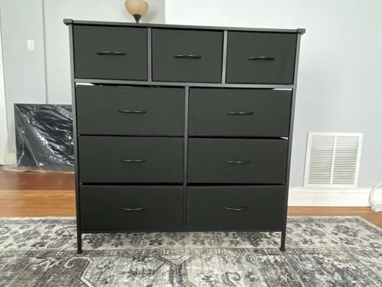 Black metal 9-drawer dresser with black fabric drawers sitting on a gray oriental rug with a gray wall and a large flat screen TV wrapped in plastic behind it