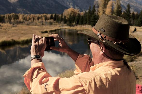 A photographer standing at a river with mirrored, calm water wearing a cowboy hat stretching out his hands clamping a small, chinese made mobile phone with which he is taking likely poor-quality images of the serene, majestic landscape while carrying a slight smirk on his face, as if he had been caught in the action of doing something unlawful, or worse. In the background the river leads towards some trees and mountains further in the back. The whole image has an orange cast to it, maybe because the sun was low, maybe because of poor post processing, maybe because orange is one of the reference colors of the Nikonians community. Hard to tell.