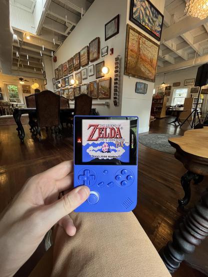 A blue Game Boy styled emulation device displays “Zelda: Links Awakeneing DX” in a bookstore filled with natural, overcast lighting. The walls of the room are adorned with paintings from a local artist. Bookshelves stand in the background on the right side of the frame.