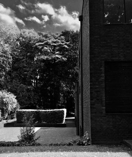 Ein Schwarz-Weiß-Bild mit einer Ecke des "Museums Haus Lange" auf der rechten Seite, umgeben von üppigem Grün und Bäumen. Eine ordentlich getrimmte Hecke erzeugt eine definierte Form im Vordergrund, mit Stufen, die zum Gebäude führen. Der Himmel ist teilweise bewölkt.
--
A black and white image featuring a corner of 'Museum Haus Lange' on the right, surrounded by lush greenery and trees. A neatly trimmed hedge creates a defined shape in the foreground, with steps leading up towards the building. The sky is partly cloudy.