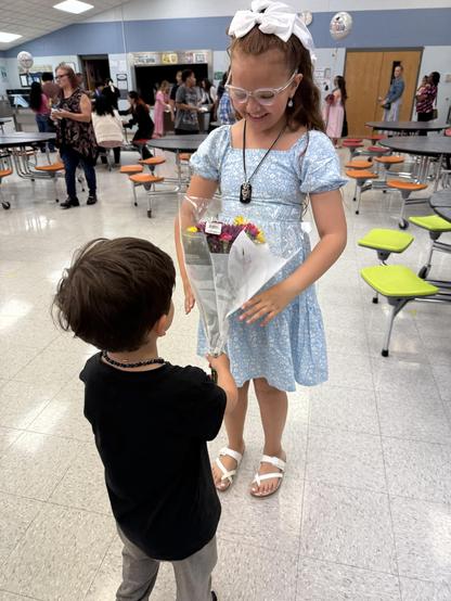 A young girl in a light blue dress and glasses is smiling while receiving a bouquet of flowers from a younger boy in a black shirt. The scene is set in a cafeteria with people in the background.