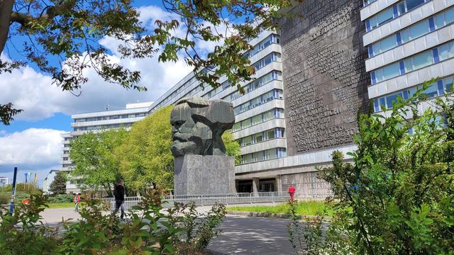 Der Karl-Marx-Kopf in Chemnitz, dahinter ein ca. 10-stöckiger Gebäuderiegel. Der Himmel ist heiter bis wolkig.