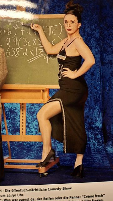 Martina Brandl steht im Abendkleid als sexy Lehrerin an einer große Schultafel und zeigt mit Brille auf der Nase und Kreide in der Hand auf eine mathematische Formel