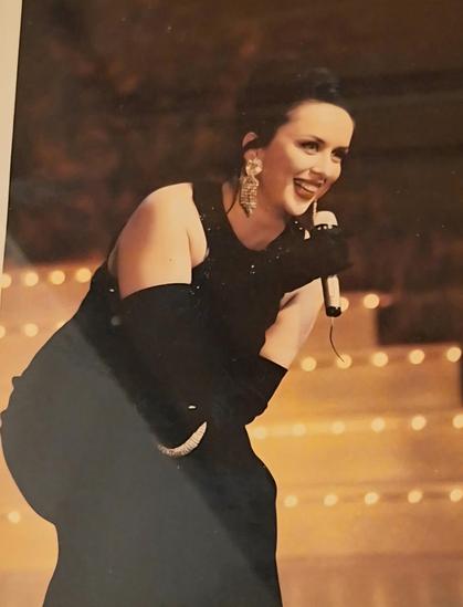 Martina Brandl im schwarzen Abendkleid mit langen schwarzen Handschuhen und Mikro in der Hand lächelnd vor einer beleuchteten Showtreppe