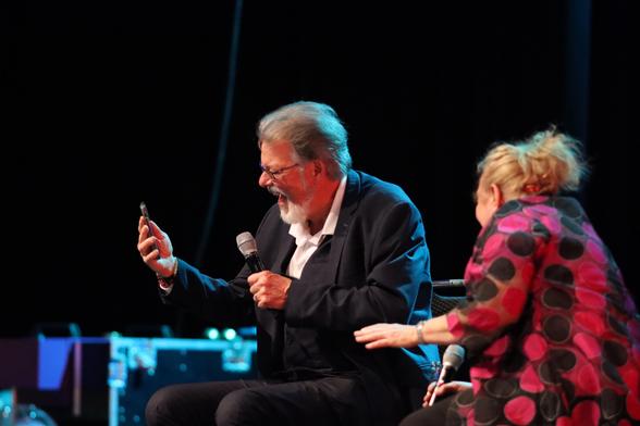A man in a suit is passionately speaking into a microphone while looking at a phone, and a woman in a polka dot top is seated next to him, laughing and engaging with him. The setting appears to be a stage or performance area.
