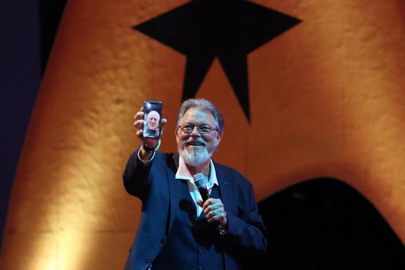 An older man with a beard is smiling and holding a smartphone up to his face, displaying a photo of another person. He is dressed in a dark suit with a white shirt and is speaking into a microphone. The background features a large star design.