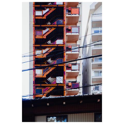 A tall apartment building's exposed emergency staircase cuts diagonally across the frame in vivid oranges and reds, its industrial rust nearly humming in the midday light. Amid the zigzag of landings and railings, a white futon is draped squarely on one level like laundry gone rogue. Behind it, pale balconies offer the more traditional drying space—but this futon has aspirations.