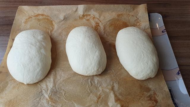 Das Bild zeigt drei Teigbällchen, die auf einem Stück Backpapier liegen. Die Teigbällchen sind weiß und weich, mit einer glatten Oberfläche und einigen kleinen Faltungen. Sie sind gleichmäßig verteilt und haben eine ovalen Form. Das Backpapierpapier ist leicht gefaltet und hat einige braune Flecken, was darauf hindeutet, dass es bereits verwendet wurde. Die Teigbällchen befinden sich auf einer Küchenarbeitsfläche. Die Teigbällchen sind wahrscheinlich vor dem Backen und werden wahrscheinlich in einem Ofen gebacken.